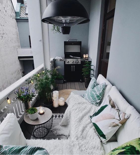 A-cozy-boho-terrace-with-a-white-sofa-bright-pillows-and-a-blanket-a-side-table-with-potted-greenery-and-blooms-plus-a-grill