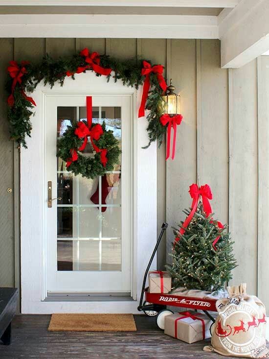 A-farmhouse-christmas-porch-with-an-evergreen-garland-with-red-bows-and-a-matching-wreath-plus-a-tree-with-lights-and-gift-boxes